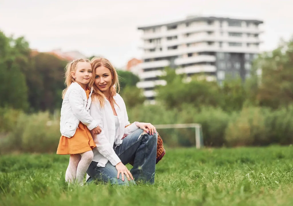 Mutter und Kind sitzen lächelnd auf einer gepflegten Grünfläche vor einem modernen Wohngebäude der Wohnbau Lörrach